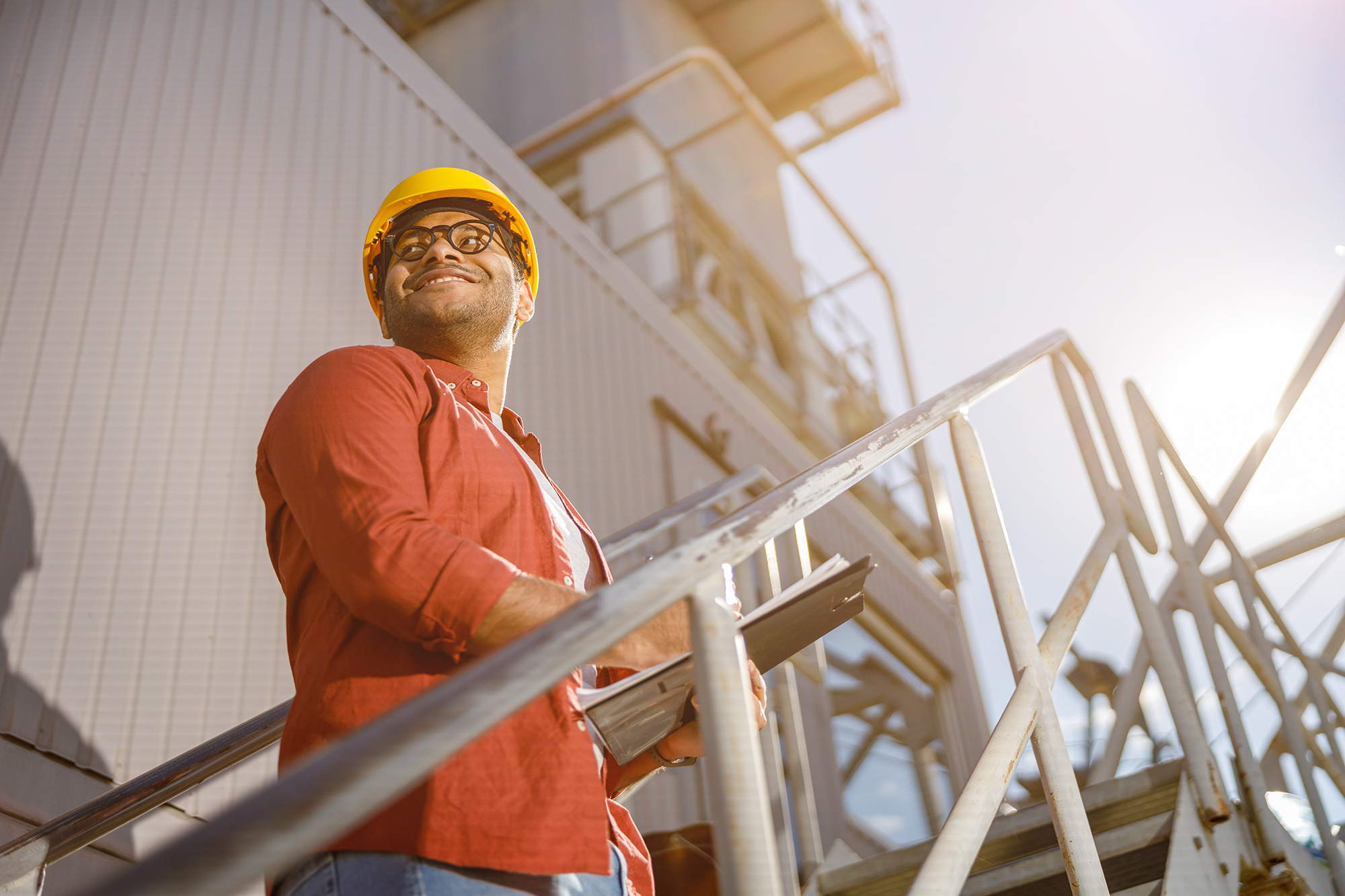 Happy male engineer wearing protective helmet and looking sideways while working at industrial zone of construction material factory